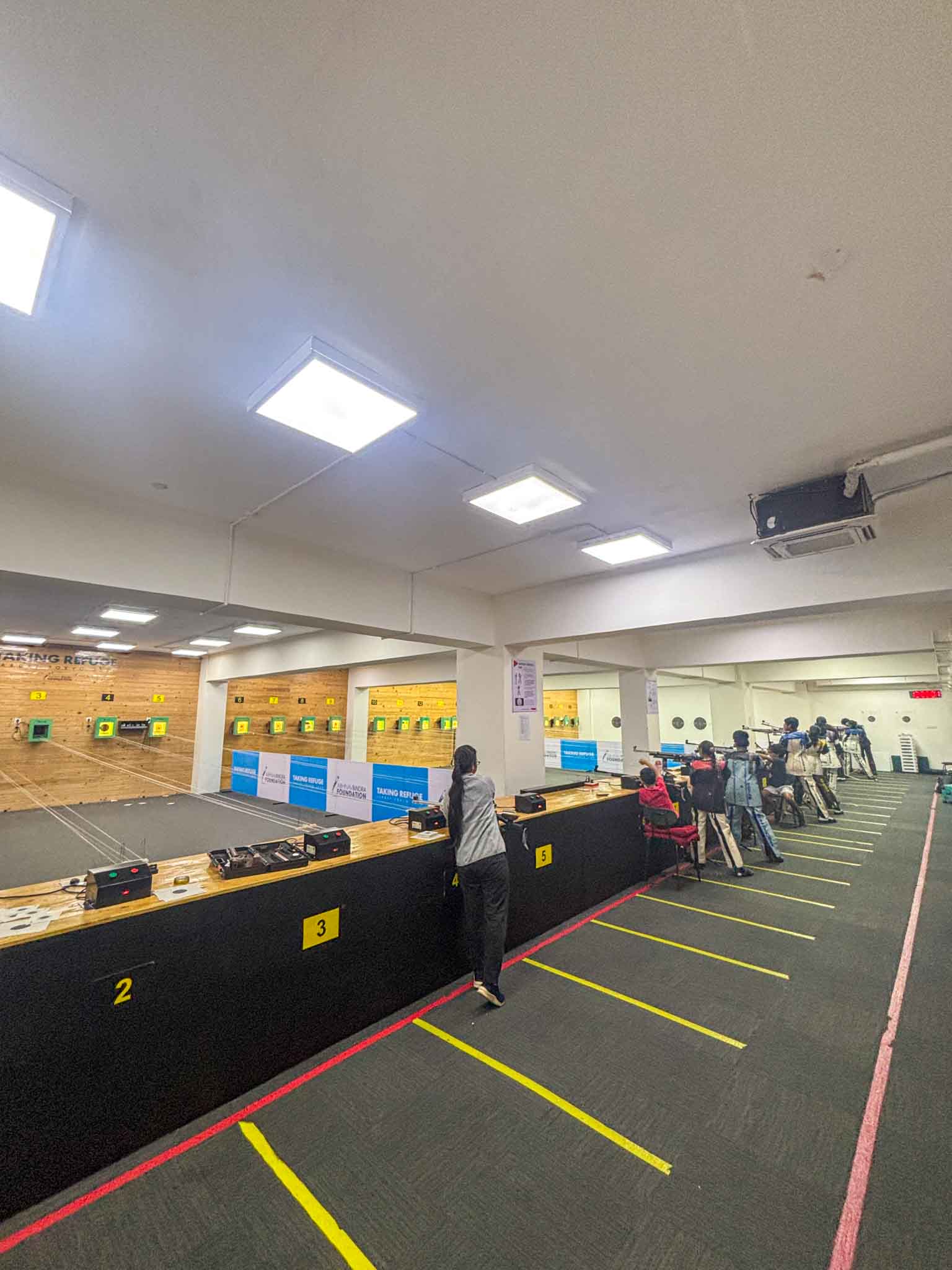 Several people practicing rifle shooting at an indoor range with marked lanes and target boards in Bengaluru.